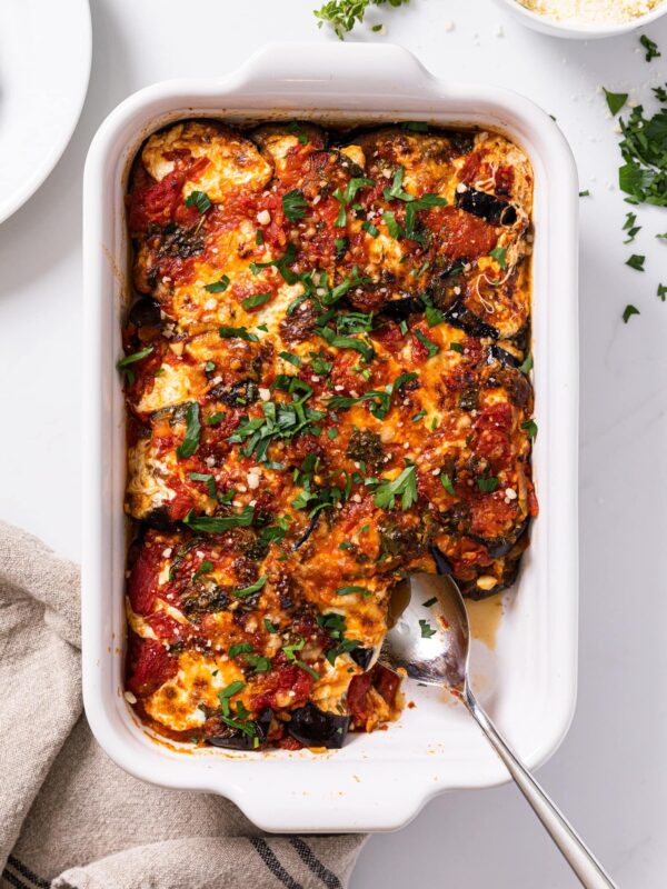 Top down view of white baking dish sitting on white countertop filled with red colored eggplant parmesan topped with sprinkling of herbs with extra cheese on the side