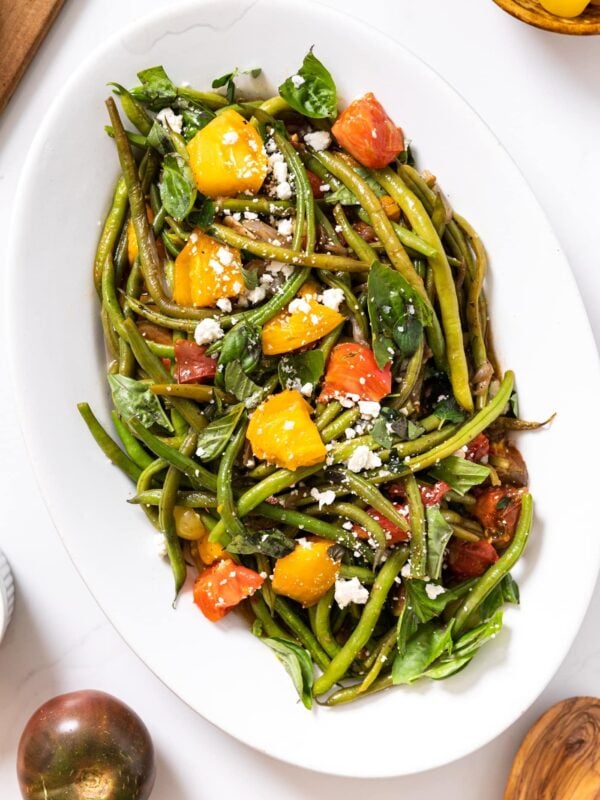 Top down view of white platter filled with green beans and roasted tomatoes sitting on white countertop