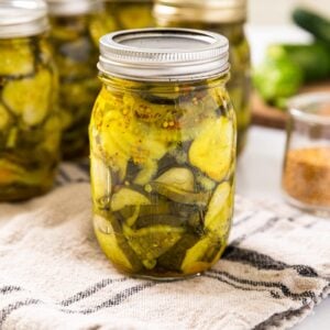 Glass jar filled with slices of pickles sitting on towel with extra glass jars in background all on white countertop