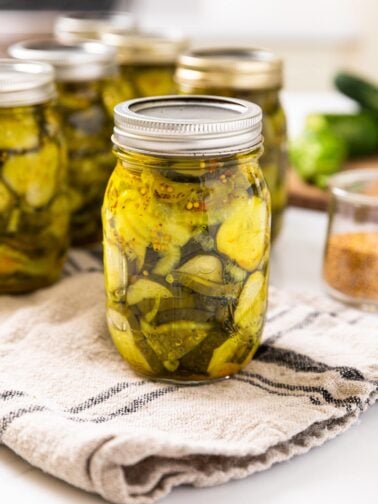 Glass jar filled with slices of pickles sitting on towel with extra glass jars in background all on white countertop
