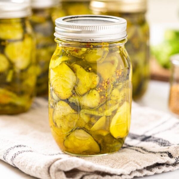 Glass jar filled with slices of green pickles in liquid sitting on towel on white countertop with other glass jars all around