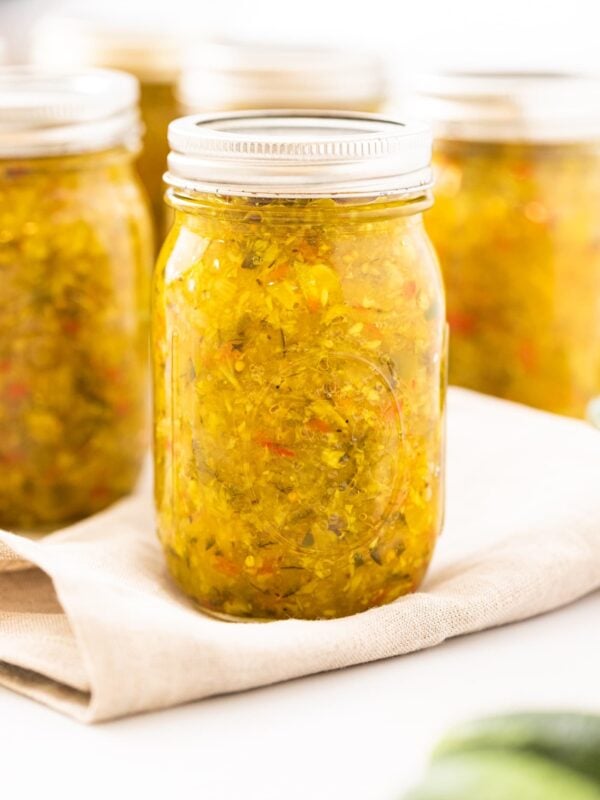 Glass canning jar filled with yellow colored pickle relish sitting on a taupe cloth with extra filled jars in background