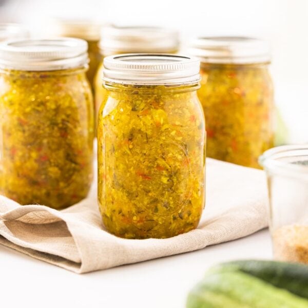 Glass canning jar sitting on white countertop filled with yellow colored pickle relish with green cucumbers in foreground