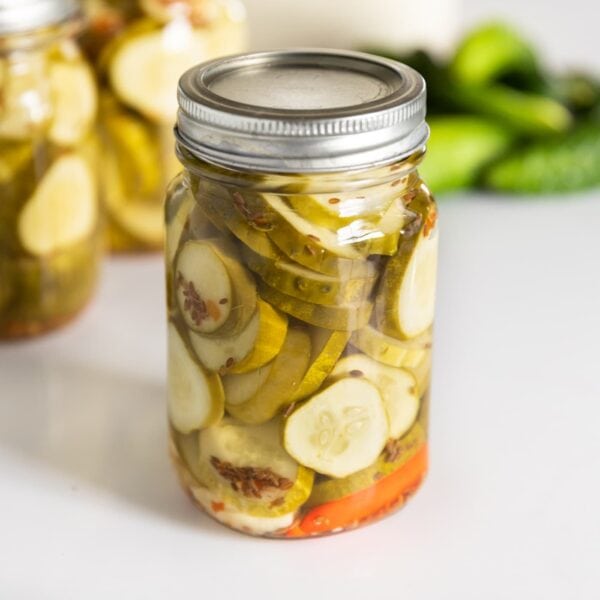 Glass jar filled with slices of spicy dill pickles sitting on white countertop with extra cucumbers in background as well as other filled canning jars