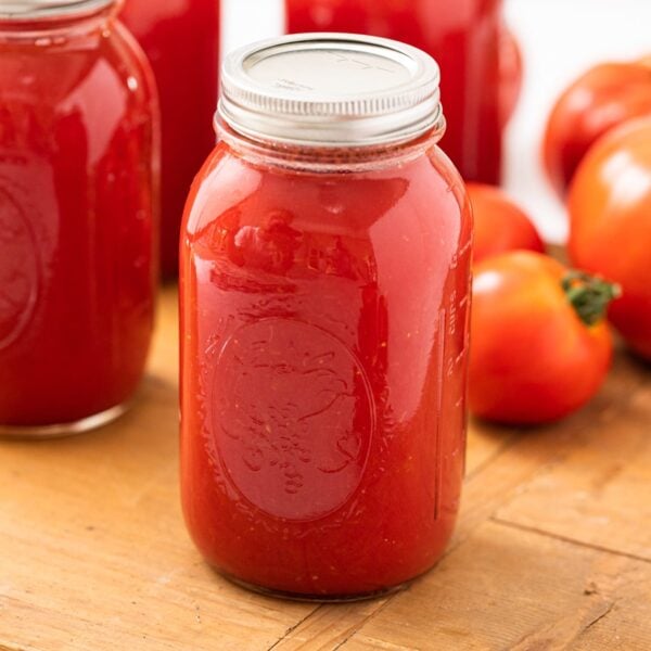 Tomato juice in glass jar with lid and ring on top sitting on wood board with unsliced red tomatoes in background along with other glass jars