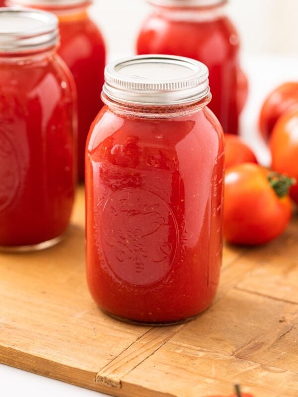 Glass jar with lid on top filled with red colored tomato juice sitting on wood board with other jars in background along with tomatoes