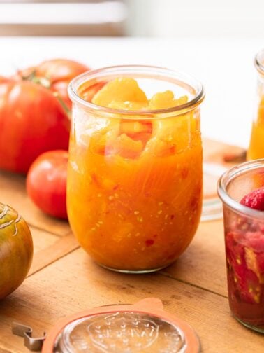 Close up view of large glass container holding yellow and orange colored tomato pieces with extra tomatoes all around all on wood board
