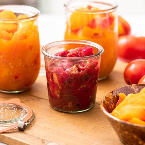 Glass containers filled with red and orange colored sliced tomatoes sitting on wood board