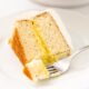 Slice of white layer cake filled with yellow pudding interior with white frosting with fork scooping off piece all on white plate