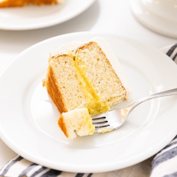 Slice of white cake with fork scooping off piece topped with white frosting and yellow colored middle layer sitting on white plate on white countertop