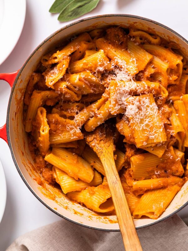 Top down view of large red pot filled with orange colored tubular pasta with wooden spoon sitting in middle all on white countertop