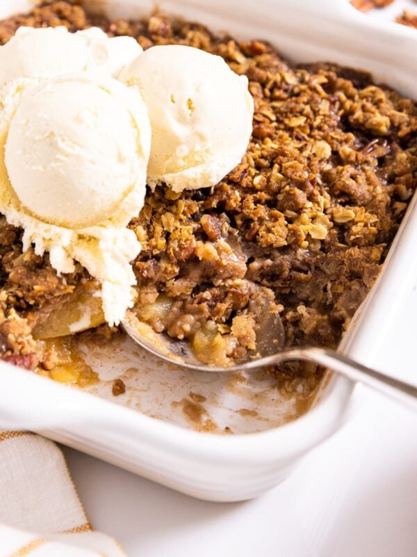 Large silver spoon sitting in white baking dish with a spoonful of apple crisp and three large scoops of ice cream on top