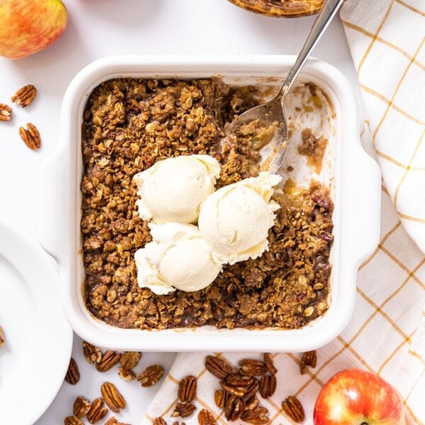 Top down view of white baking dish filled with brown colored apple crisp with three scoops of vanilla ice cream on top all sitting on white countertop with baking ingredients all around