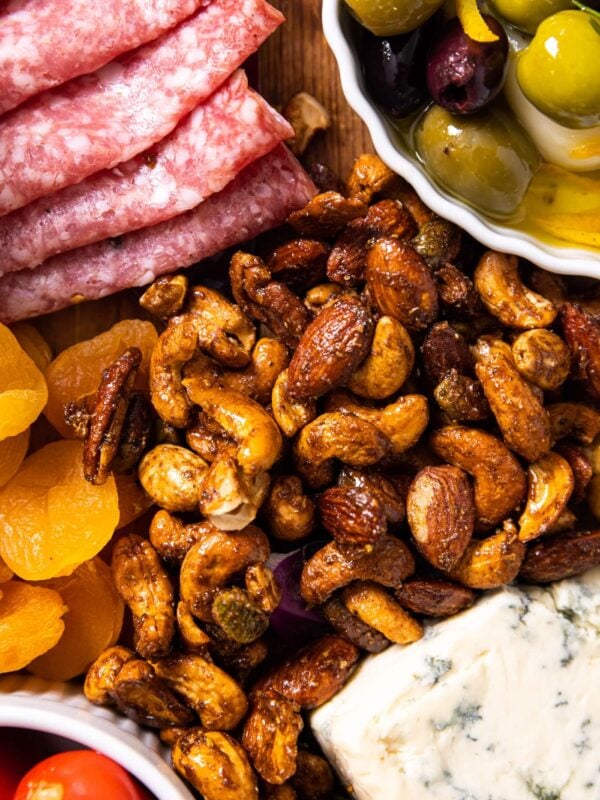 Top down view of spiced nut mixture sitting on wood board amongst olives, meats, dried fruit, and cheese