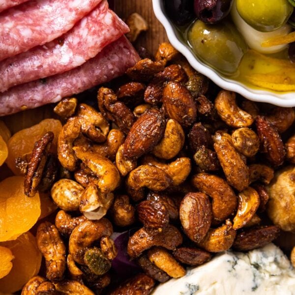 Top down view of a fall spiced nut mix sitting on wood board surrounded by dried fruits, cheese, and meats