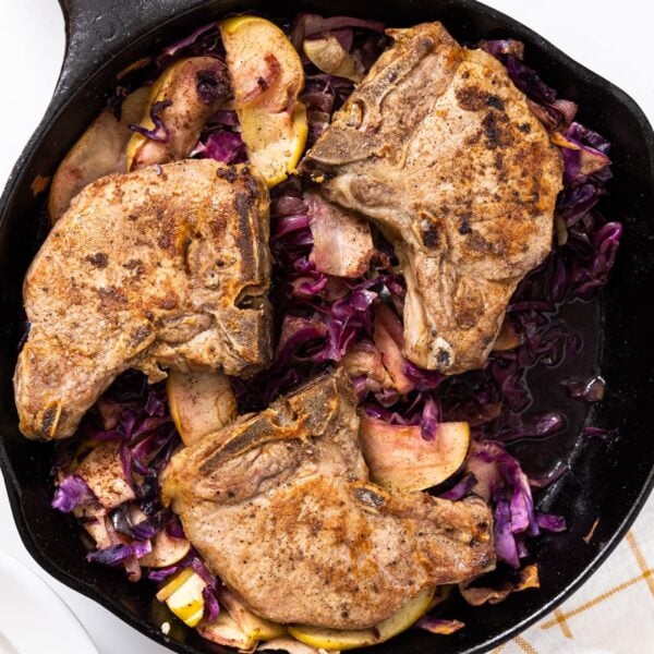 Top down view of black cast iron skillet sitting on white countertop with pork chops in the pan along with cabbage and apples