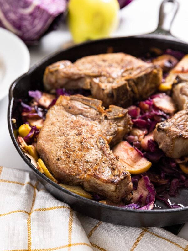 Large black skillet filled with browned pork chop with red cabbage underneath with yellow checked cloth underneath pan