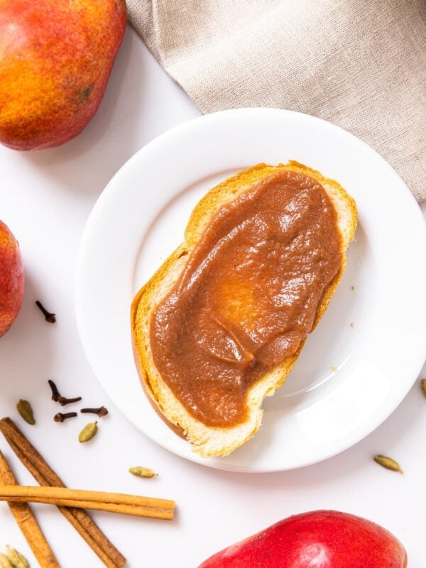Top down view of white plate sitting on white countertop with slice of bread topped with brown colored pear butter and pears and spices on the countertop
