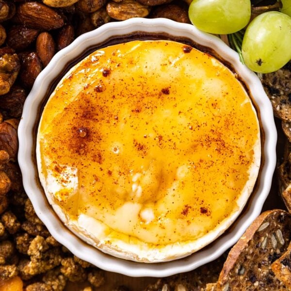 Top down view of small white bowl sitting amongst crackers and spiced nuts filled with golden colored baked Brie with chipotle powder on top