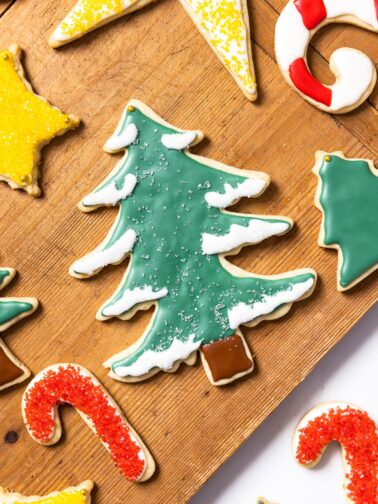 Top down view of wood cutting board sitting on white countertop with decorated sugar cookies on top in all kinds of colors