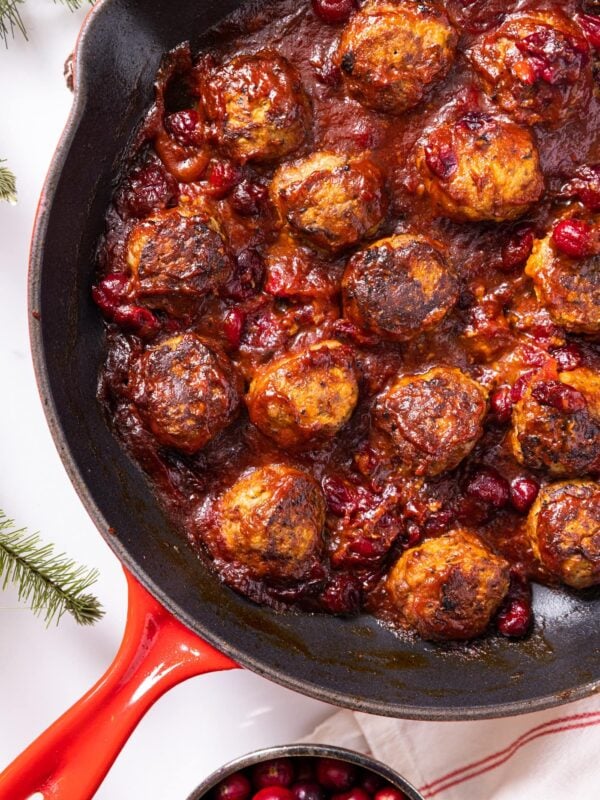 Top down view of red skillet sitting on white countertop filled with small meatballs covered in dark maroon sauce made from cranberries