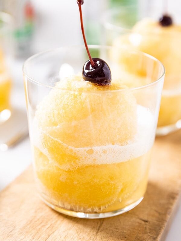 Clear glass sitting on wood board with yellow colored pineapple slush scooped inside with soda fizzing on top with a cherry on top as well