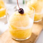 Clear glass sitting on wood board with pineapple slush sitting inside with cherry on top all on white countertop