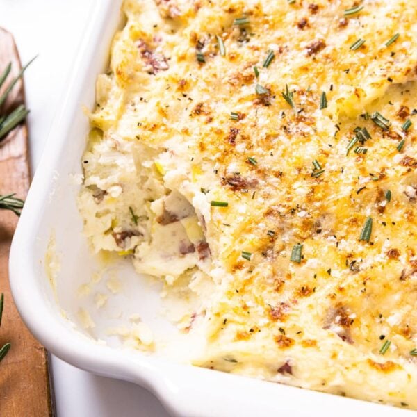 White baking dish sitting on white countertop filled with smashed potatoes with golden brown top with wood cutting board with rosemary sitting on top