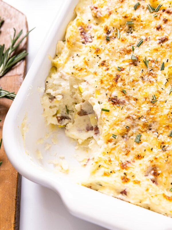 White baking dish filled with smashed potatoes with golden brown top with brown cutting board sitting beside with rosemary on top all on white countertop
