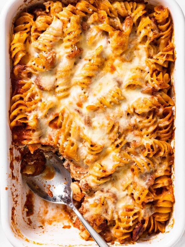 Top down view of white baking dish filled with pasta covered in red pasta sauce with cheese sprinkled over the top with spoon taking scoop out