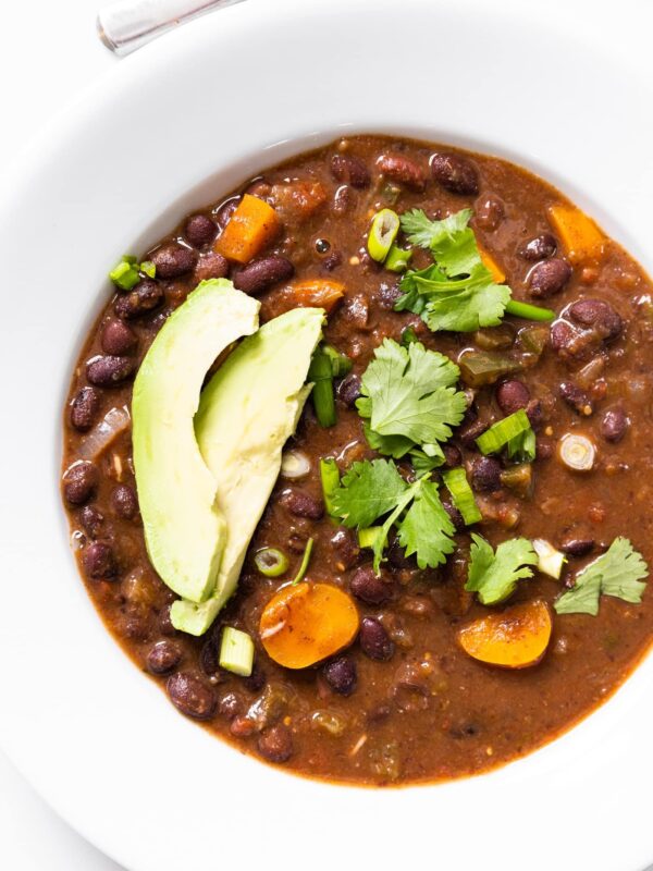 Top down view of white bowl filled with black bean soup with pieces of carrot and slices of avocado and cilantro on top with spoon at top of frame