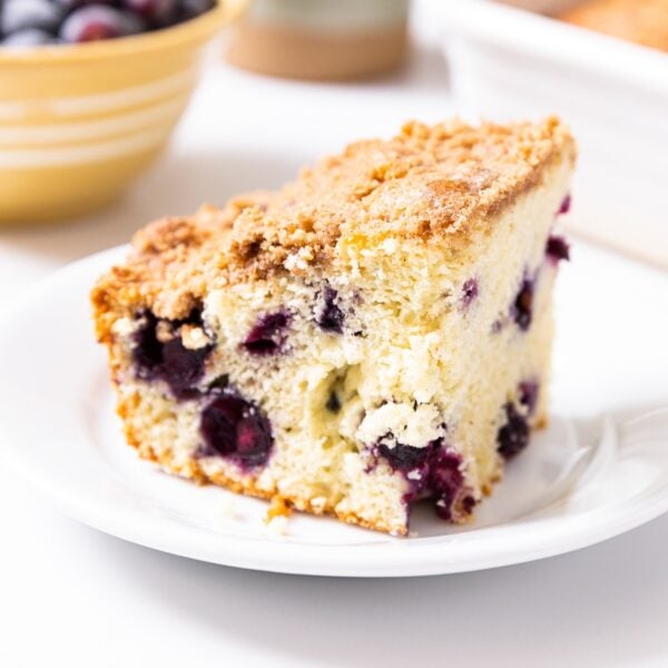 Slice of blueberry coffee cake sitting on white plate with bowl of blueberries in background and rest of coffee cake in pan in background all on white countertop