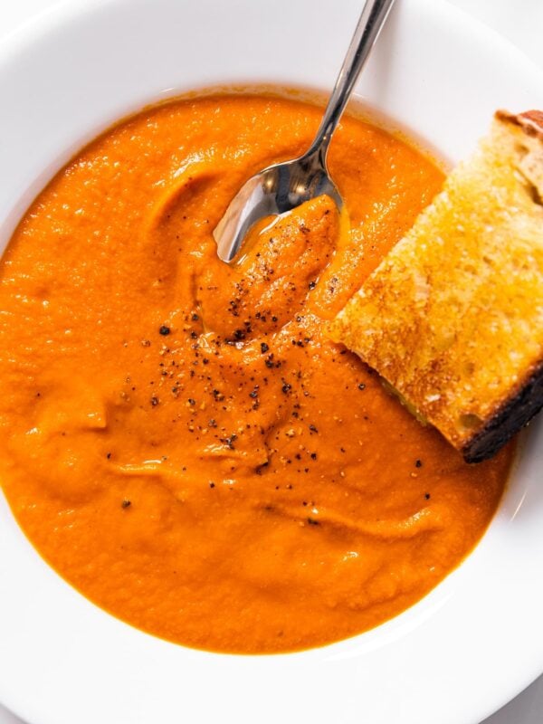 Top down view of white bowl filled with red colored creamy tomato soup with pepper on top and spoon scooping out along with slice of grilled cheese sandwich