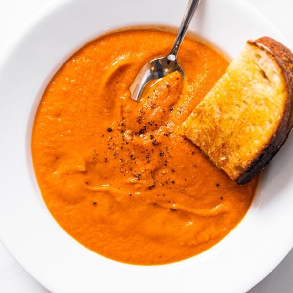 Top down view of white bowl filled with red colored creamy tomato soup with slice of grilled cheese sandwich sitting on side of bowl all on white countertop