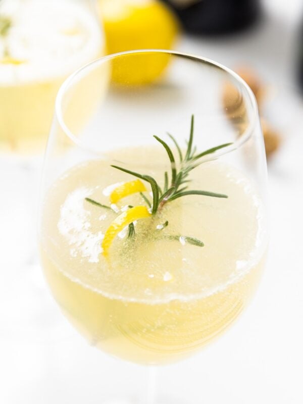 Wine glass filled with yellow colored French 75 cocktail with sprig of rosemary and lemon peel twist sitting on white countertop