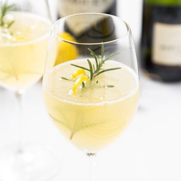 Wine glass filled with French 75 cocktail with sprig of rosemary and lemon peel sitting on white countertop with rest of ingredients in background