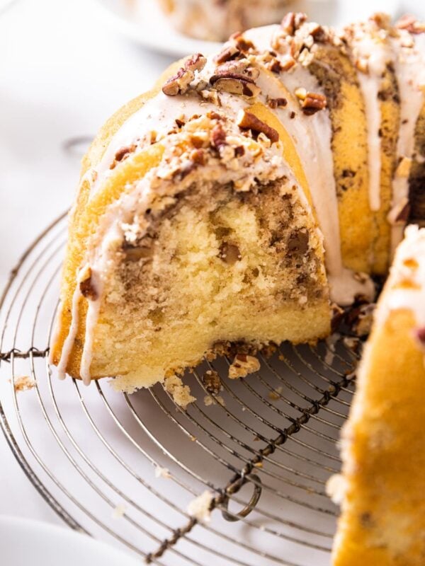 Side view of yellow coffee cake with white glaze on top with piece missing from cake all on wire circular rack sitting on white countertop