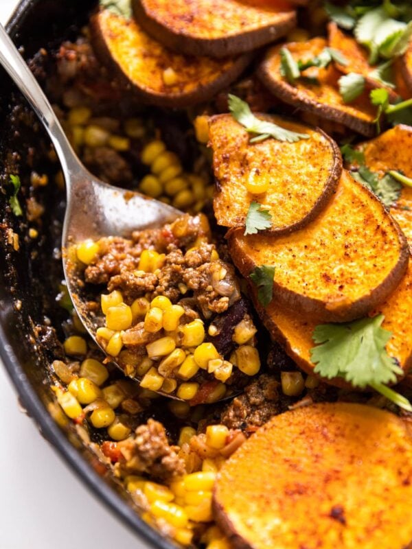 Serving spoon with scoop of taco bake sitting in black skillet with pieces of sweet potato all around on white countertop