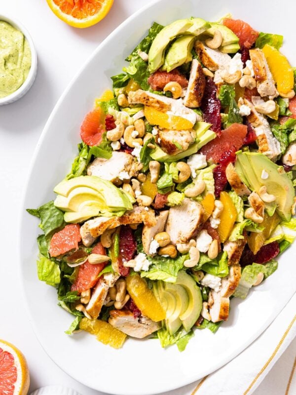 Top down view of white serving platter on white countertop filled with citrus chicken salad topped with green dressing and cashews with excess citrus all around