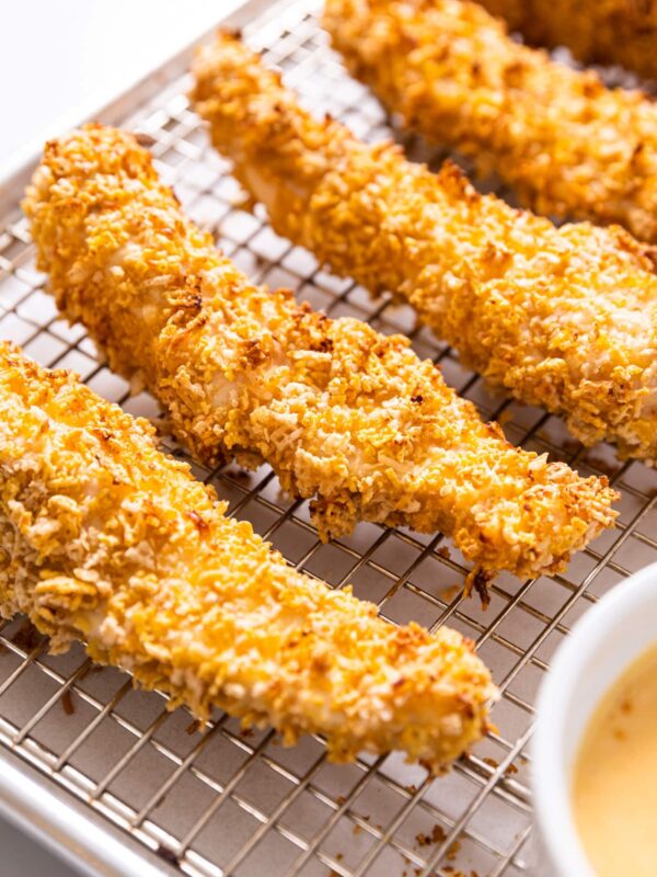 Long strip of baked crispy chicken strip sitting on metal cooling rack with small bowl filled with honey mustard in front