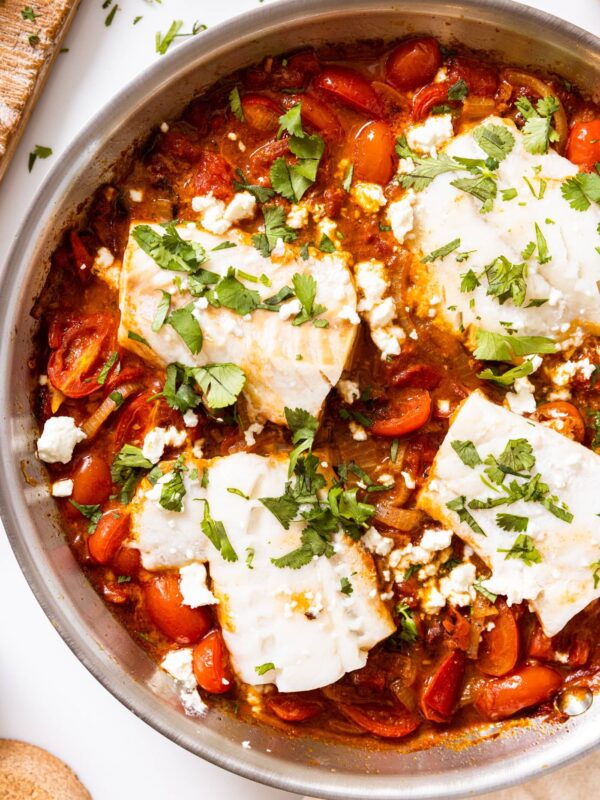 Top down view of metal skillet sitting on white countertop filled with red sauce with white filets of cod topped with chopped cilantro