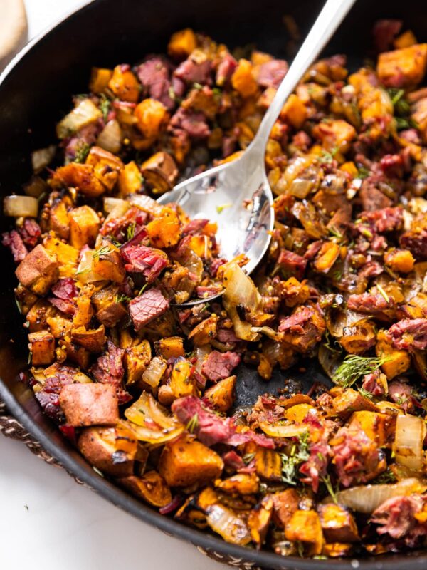 Large black skillet filled with corned beef and sweet potato hash after being cooked on the stove with spoon sitting in skillet about to scoop a serving