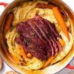 Top down view of red Dutch oven filled with yellow cabbage and carrots and red colored corned beef all on white countertop