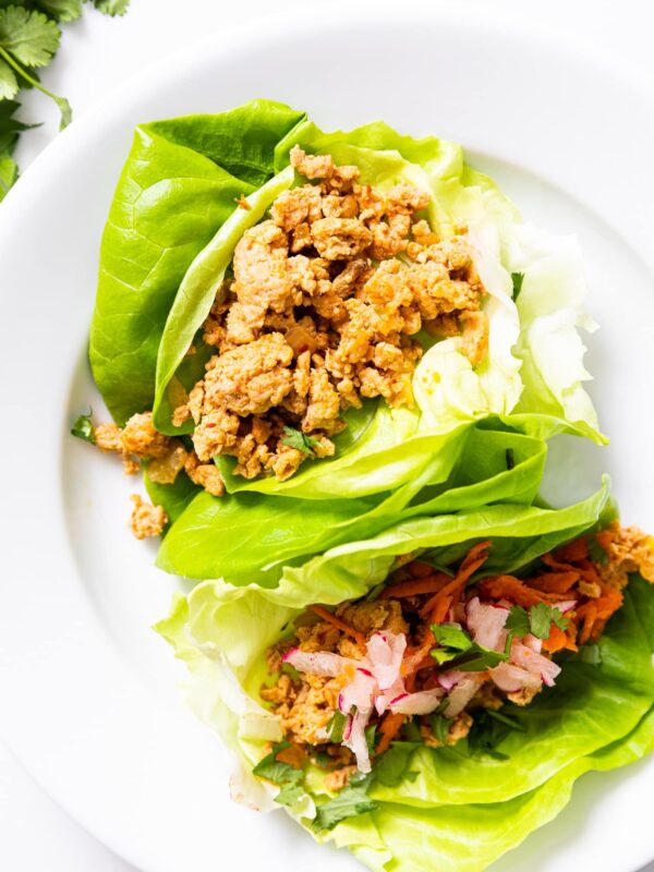 Top down view of two chicken lettuce cups sitting on white plate with toppings on one cup