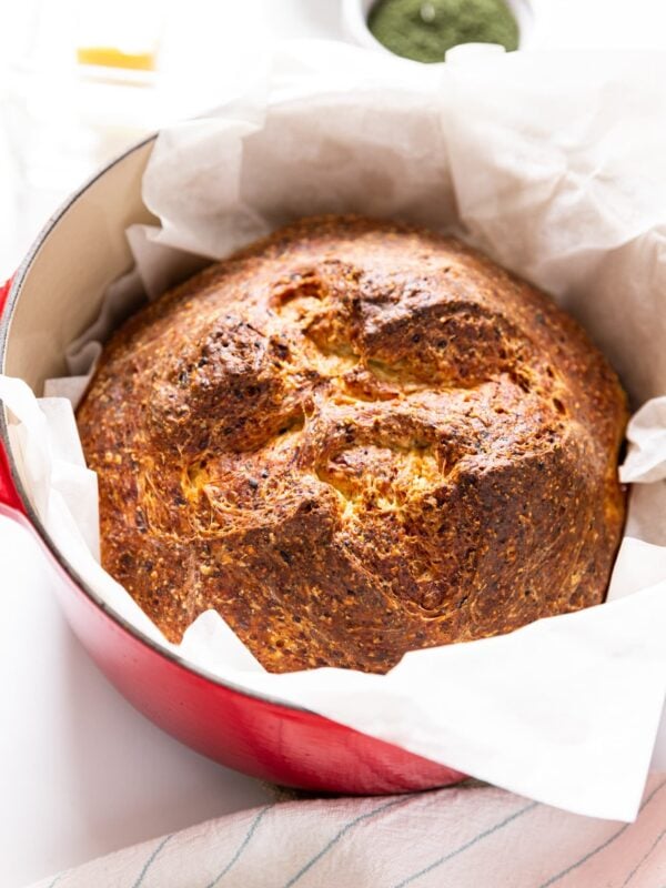 Dilly bread with golden brown top sitting on piece of white parchment scrunched into red Dutch oven sitting on white countertop