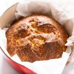 Large circular dilly bread loaf with golden top sitting in red Dutch oven with scrunched piece of parchment paper underneath
