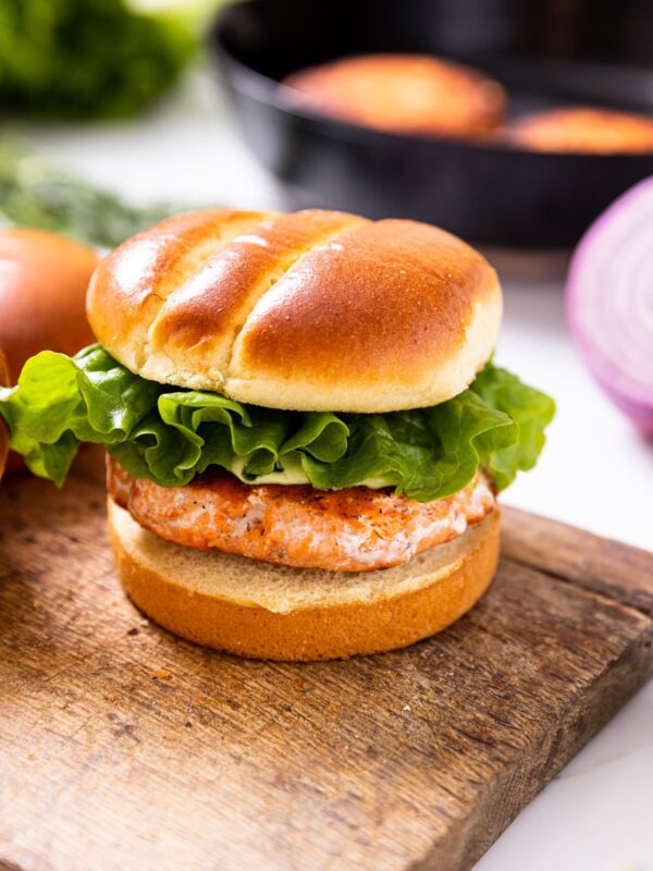 Large salmon burger slathered with dill aioli sitting between brioche buns on wood board with black skillet in background