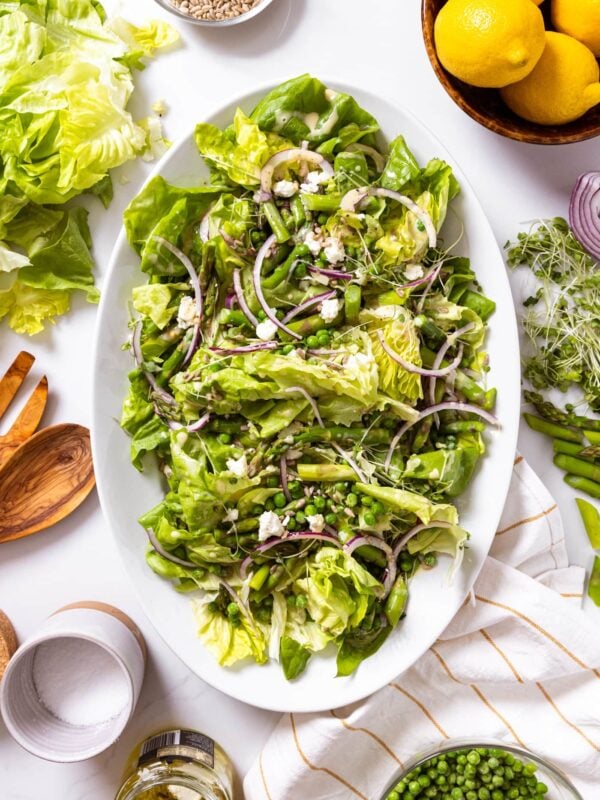 Top down view of white serving platter with lots of lettuce topped with red onion and asparagus and peas with bowl of lemons beside along with serving tongs and other salad components