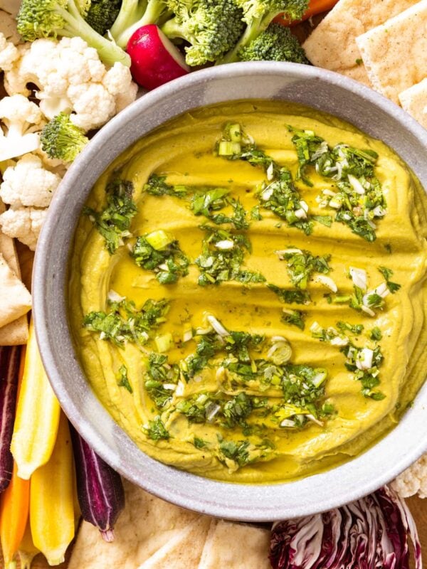 Top down view of green colored hummus topped with cilantro and olive oil sitting in white serving bowl with vegetables and pita all around