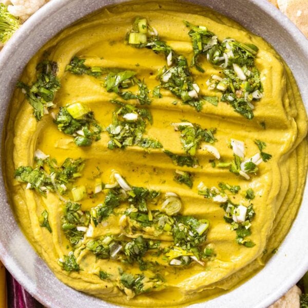 Top down view of white pottery serving bowl sitting on wood board with vegetables all around as well as pita with green colored hummus with cilantro topping sitting in the bowl